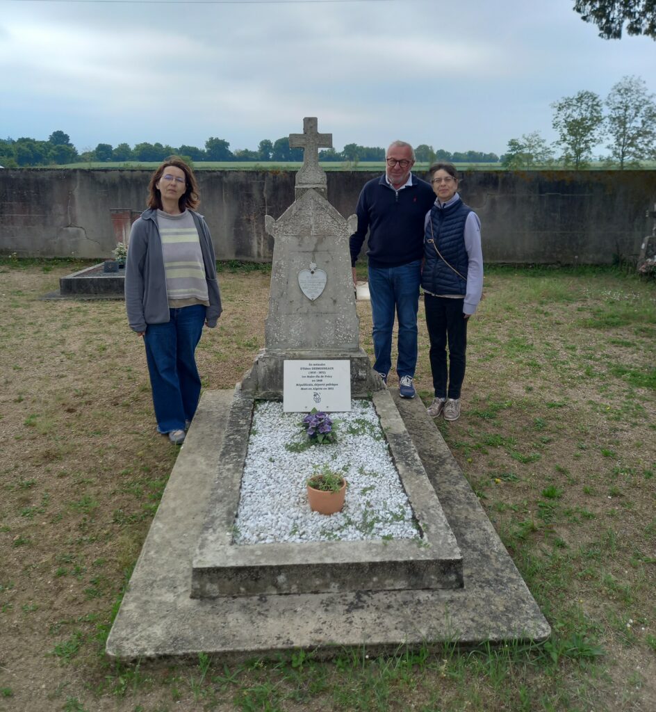 Three adults pose beside a stone cross memorial in a graveyard, with a low wall and open fields in the background.