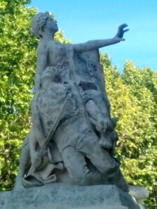 Marble statue of a seated figure with an outstretched right arm, pedestal bearing partial inscription, set against green trees and blue sky.