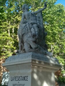 Stone statue of a seated man with an outstretched arm, atop a pedestal, surrounded by trees; inscription begins with 'LA RESISTANCE' on the pedestal.