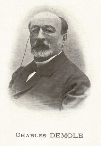 Portrait of Charles Demole, an elderly man with a white beard and round glasses, wearing a dark suit.