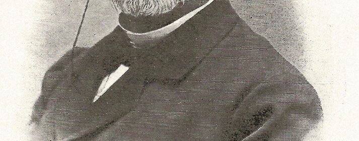 Portrait of Charles Demole, an elderly man with a white beard and round glasses, wearing a dark suit.