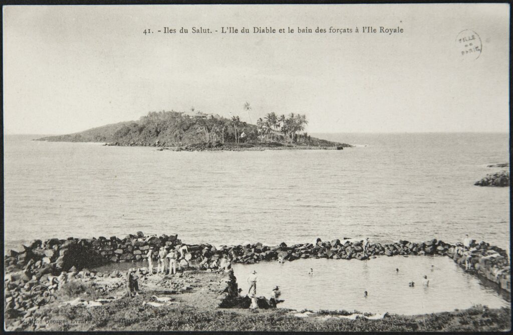 Coastal scene with a rocky seawall in the foreground and a small palm-covered island in the calm sea; crowd gathered along the shore.