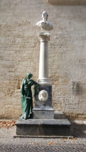 White bust on a tall marble column with a draped base sculpture, set against a textured stone wall; leaves on the ground nearby.