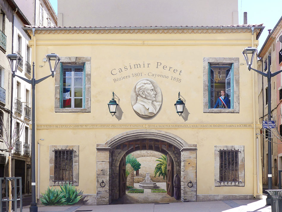 Yellow historic building with 'Casimir Peret' inscription, two windows, and a central arched gateway to a courtyard.
